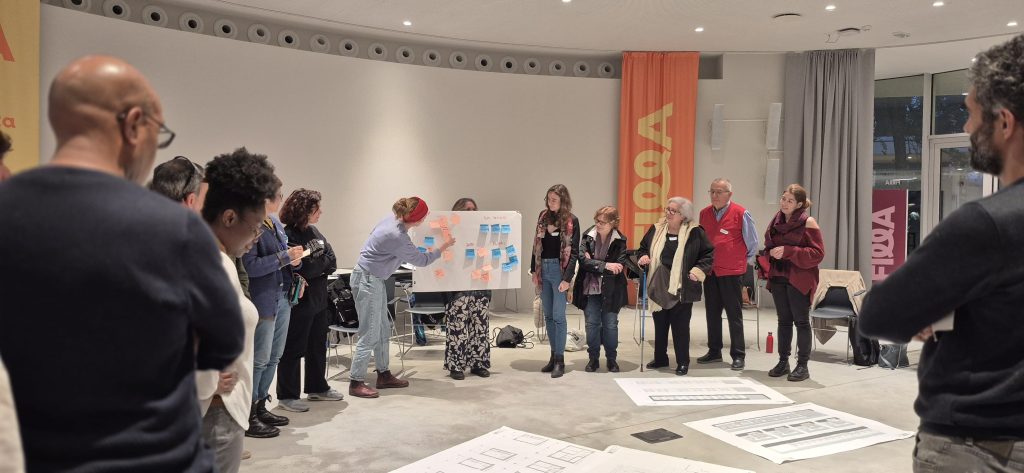 I partecipanti stanno in piedi, disposti a cerchio, mentre una di loro incolla un post-it su un cartellone